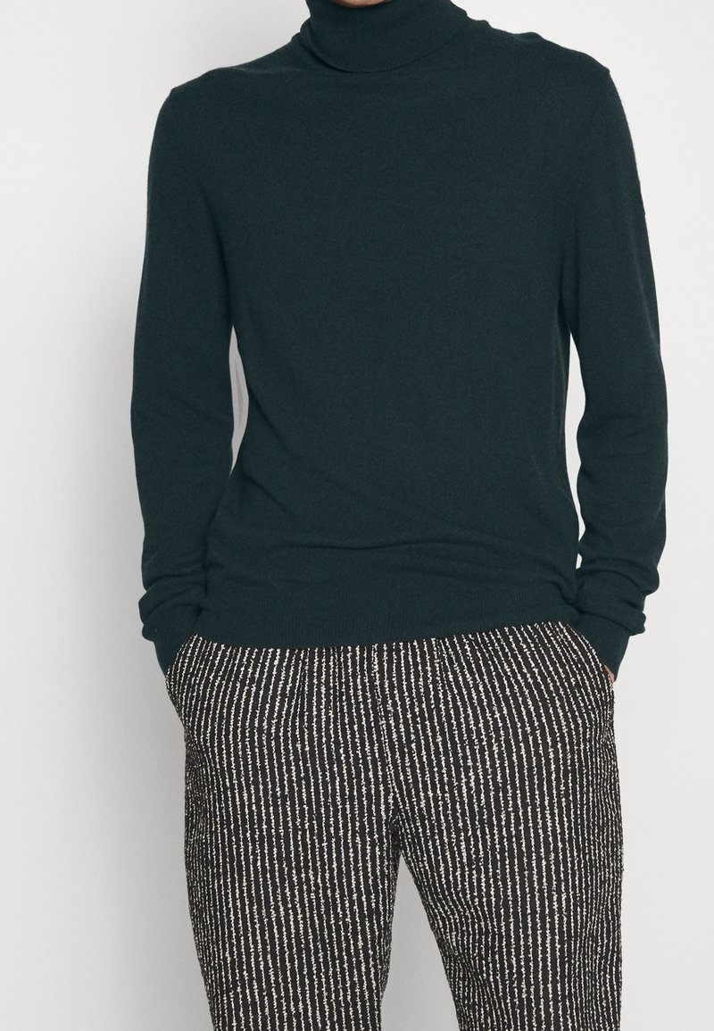Personne portant un pull à col roulé vert foncé à manches longues et un pantalon à rayures verticales noires et blanches, les mains dans les poches.