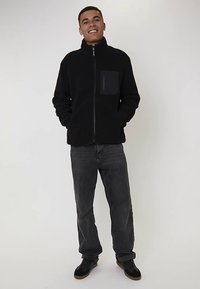 Czarna polarowa kurtka ze stójką i zamkiem z przodu, z kontrastującą ciemną kieszenią. Noszona z szarymi dżinsami i czarnymi butami.