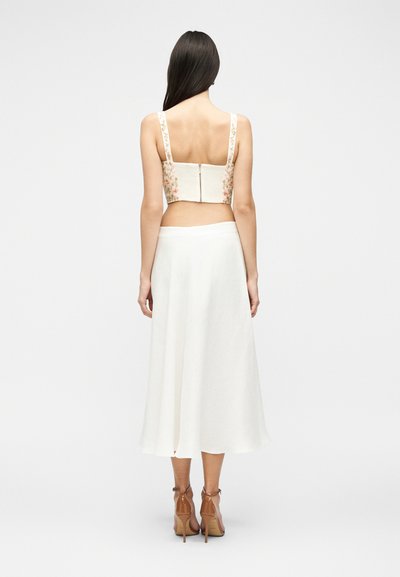 Γυναίκα που στέκεται με την πλάτη προς τα εμάς, φοράει ένα αμάνικο φλοράλ τοπ κομμένο, λευκή midi φούστα και ταμπά σανδάλια με ψηλό τακούνι σε απλό φόντο.