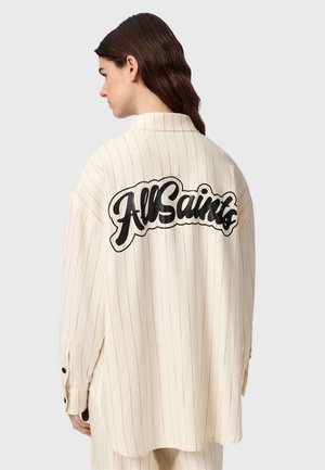 Vrouw met bruin haar die een loszittend crèmekleurig gestreept overhemd draagt met "AllSaints" in grote zwarte letters op de achterkant, met haar rug naar de kijker toe.