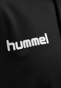 Schwarzer Stoff mit einer glatten Textur, der ein auffälliges weißes "hummel"-Logo in einer abgerundeten serifenlosen Schriftart zeigt.
