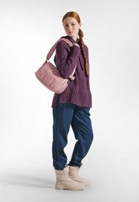 Borsa a spalla imbottita rosa con un accento arricciato, appesa a un modello che indossa un maglione viola scuro e jeans blu, completati da stivaletti beige.