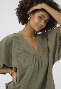 Olívazöld blúz V-kivágással, laza ujjakkal és pliszézett dizájnnal. Virágos hímzés díszíti a nyakkivágást. Puha, texturált anyag.