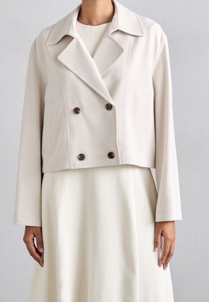 Kobieta ubrana w beżową, krótką, dwurzędową marynarkę z czterema brązowymi guzikami, nałożoną na dopasowaną długą sukienkę, na neutralnym tle.