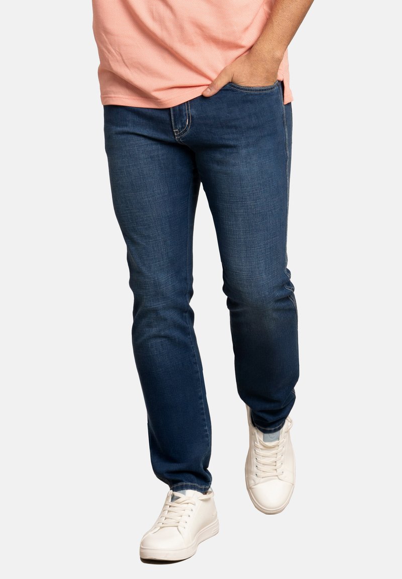 Jeans de mezclilla azul oscuro con un ajuste slim, que presentan un diseño clásico de cinco bolsillos y costuras sutiles. Combinados con zapatillas blancas.