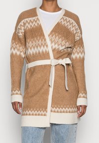 Beige stickad kappa med krämfärgade accenter, med ett geometriskt mönster, öppen fram och ett knytbelte i midjan. Ribbade ärmslut och nederkant.