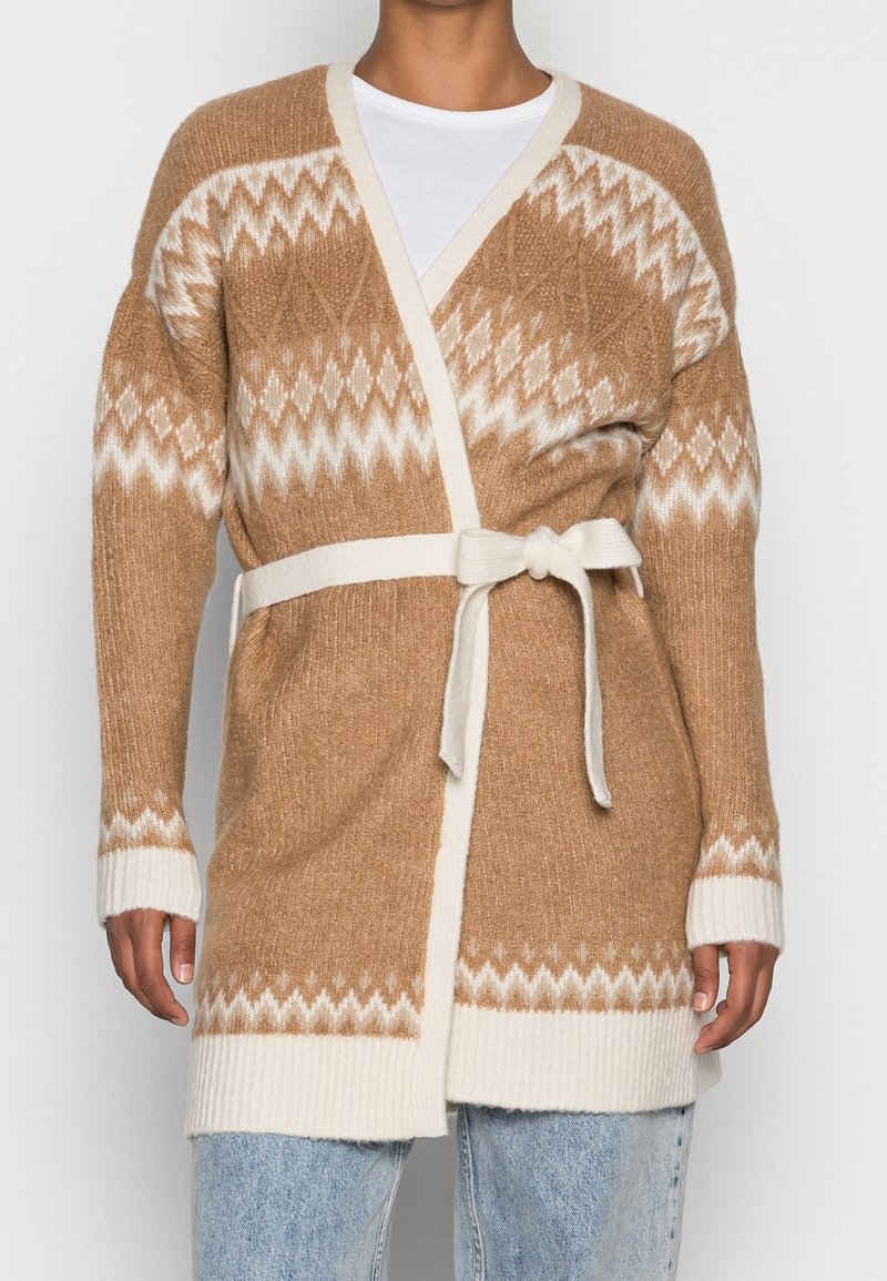 Beige stickad kappa med krämfärgade accenter, med ett geometriskt mönster, öppen fram och ett knytbelte i midjan. Ribbade ärmslut och nederkant.