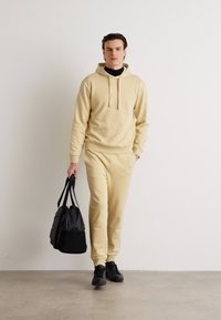 Hanorac beige și pantaloni de trening asortați, dintr-un material moale, având glugă cu șnur și buzunare laterale, asortați cu adidași negri și o geantă.