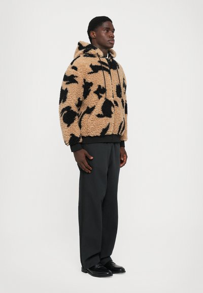 Man die naar rechts kijkt en een beige en zwart geblokte fluffy capuchonjas draagt, donkere wijde broek en zwarte nette schoenen.