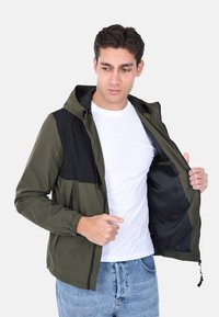 Giacca con cappuccio in verde oliva e nero, con chiusura a zip frontale, polsini elastici e foderata interna liscia. Esposta aperta con tasche.