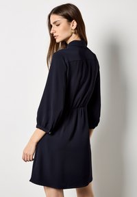 Dunkelblaues Kleid mit Kragen, Dreiviertelärmeln und elastischem Tailleband. Glatte Textur, über dem Knie endende Länge, einfaches Design.