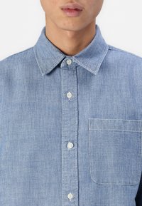 Lyseblå denimskjorte med klassisk krave, forlommer, knaplukning og subtil tekstur. Har hvide knapper og syningdetaljer.