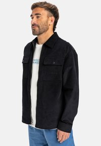 Giacca in cotone a coste nera con zip frontale, due tasche sul petto e un branding sottile. Tessuto morbido con una vestibilità strutturata.