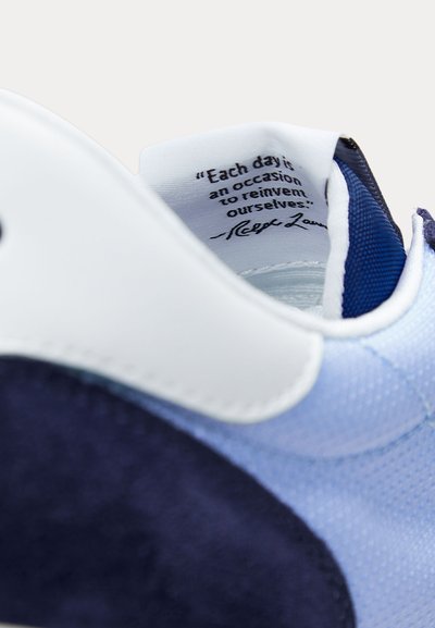 Balta un zila sporta apavu ar redzamu iekšējās mēles tekstu: "Katru dienu ir iespēja sevi pārveidot", parakstīts Ralph Lauren.