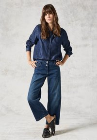 Camicia a righe navy con bottoni, pantaloni larghi in denim scuro, e zoccoli neri con borchie dorate. Sfondo chiaro e strutturato.