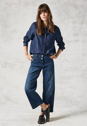 Camicia a righe navy con bottoni, pantaloni larghi in denim scuro, e zoccoli neri con borchie dorate. Sfondo chiaro e strutturato.