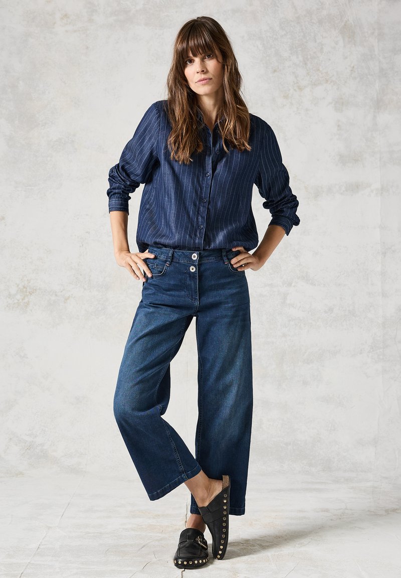 Camicia a righe navy con bottoni, pantaloni larghi in denim scuro, e zoccoli neri con borchie dorate. Sfondo chiaro e strutturato.