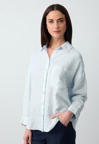 Lichtblauwe linnen shirt met een knoopsluiting aan de voorkant, een enkele borstzak en lange mouwen. Zachte textuur met een relaxte pasvorm en opgerolde manchetten.