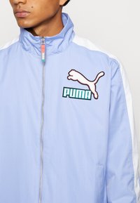 Lyseblå jakke med lynlås, der har hvide sidelapper, ribbet tekstur, broderet Puma-logo og en funktionel lynlås med farverig trækstrop.