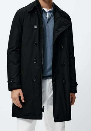 Homme portant un manteau croisé noir sur un polo bleu et un pantalon blanc, debout devant un fond uni.