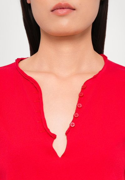Red cotton top featuring a shallow V-neck, raw edge, and seven buttons down the front. Smooth texture with subtle stitching detail.