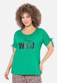 Groen T-shirt met korte mouwen en een ronde hals, met zwarte letters "Stay WILD" en geplooide mouwen met strikjes.