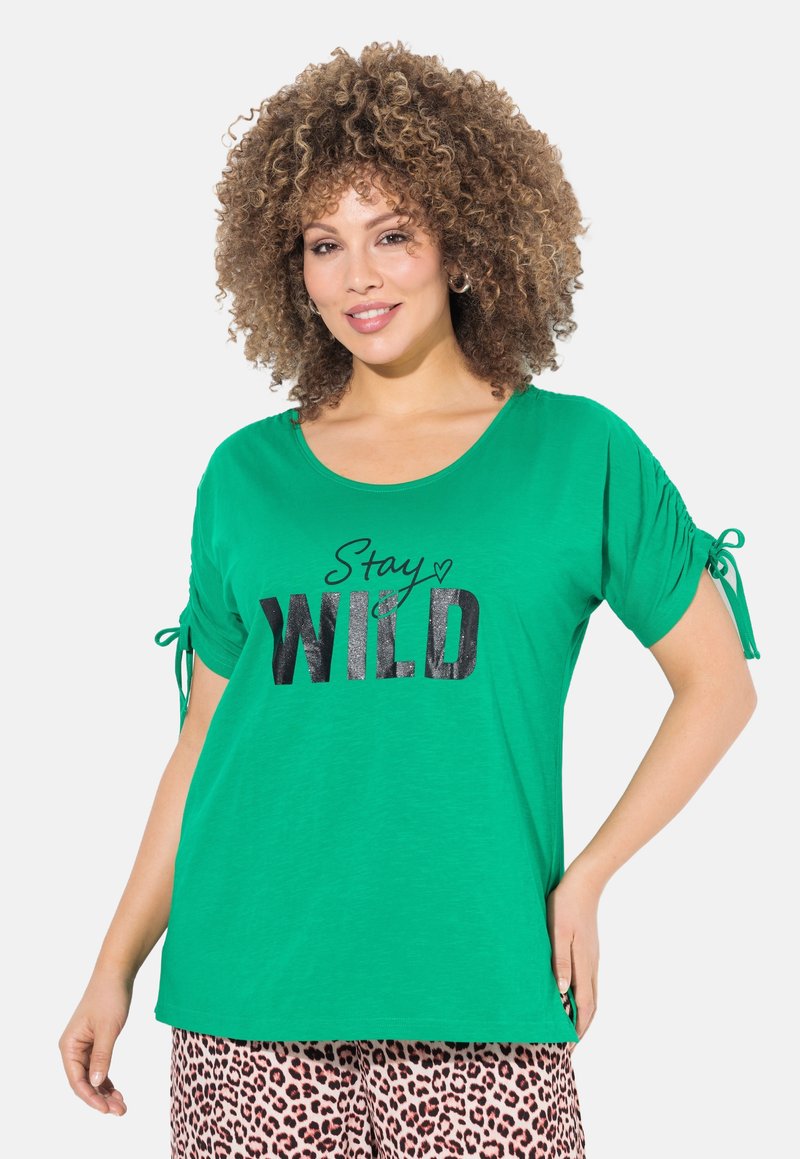 Groen T-shirt met korte mouwen en een ronde hals, met zwarte letters "Stay WILD" en geplooide mouwen met strikjes.