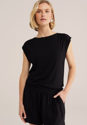 Blonde vrouw die een zwarte mouwloze top en zwarte korte broek met elastische tailleband draagt, poseert met één hand in de zak tegen een effen achtergrond.