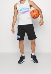 Biały top bez rękawów z kolorową grafiką; czarne szorty z nadrukowanym logo; kosz do koszykówki w ręku; czarne buty sportowe z białymi akcentami.