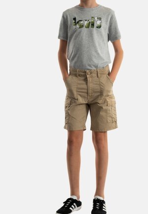 Barn iført grå Levi's t-shirt, beige cargoshorts med lommer, sort-hvide sneakers, står med hænderne i lommerne mod hvid baggrund.