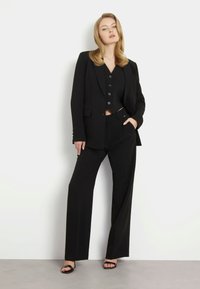 Crni odijelo po mjeri sastoji se od strukturirane sako jakne, dugog prsluka i hlača širokih nogavica. Materijal izgleda glatko s elegantnim, profesionalnim dizajnom.