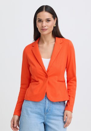 Frau mit langen dunklen Haaren, die goldene Ohrringe, ein oranges Blazer über einem weißen Oberteil und hellblaue Jeans trägt, steht vor einem weißen Hintergrund.