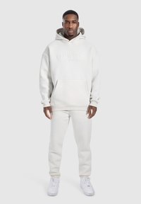 Hellgrauer Hoodie und passende Jogginghose aus weichem, strukturiertem Stoff. Der Hoodie verfügt über eine Fronttasche und geprägte Logo-Details.