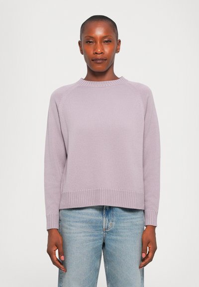 Donna che indossa un maglione a maglia viola chiaro e jeans blu chiaro, in piedi contro uno sfondo bianco semplice.