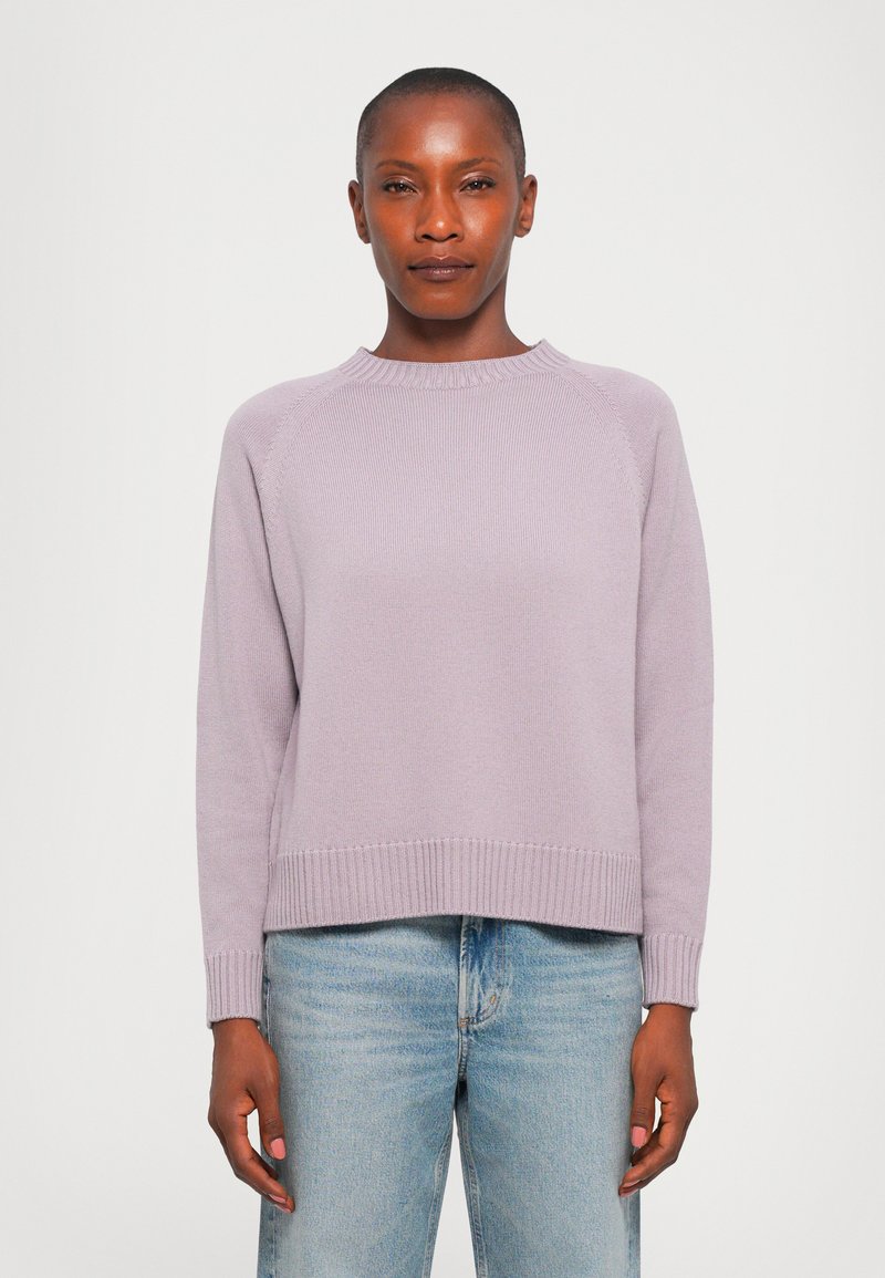 Woman wearing a light purple knit sweater and light blue jeans, standing against a plain white background.
