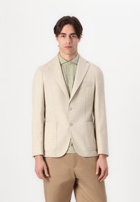 Blazer beige texturé avec revers pointus, deux boutons et poches à rabat, porté sur une chemise col vert clair et un pantalon beige.
