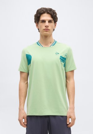 Νέος με κοντά σγουρά μαλλιά που φοράει ανοιχτό πράσινο κοντομάνικο πουκάμισο με τυρκουάζ γεωμετρικά σχέδια και σκούρα σορτς, στέκεται μπροστά σε απλό φόντο.