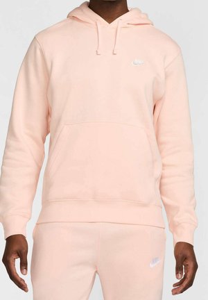 Mężczyzna ubrany w jasnoróżową bluzę z kapturem Nike oraz dopasowane spodnie dresowe z białymi logo Nike na klatce piersiowej i udzie.