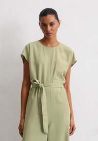 Salopette verde oliva con maniche corte, una tasca frontale e una cintura annodata. Il tessuto morbido e la vestibilità comoda creano un design minimalista.