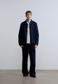 Μαύρο μπουφάν με φερμουάρ πάνω από λευκό t-shirt, συνδυασμένο με φαρδιά μαύρη παντελόνια και μαύρα παπούτσια. Απλός, μοντέρνος σχεδιασμός και ουδέτερα χρώματα.