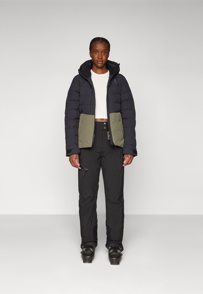 Daunenjacke mit schwarzem Obenteil und olivgrünem Unterteil. Verfügt über eine Kapuze und Reißverschluss-Taschen. Kombiniert mit schwarzen Hosen und Skischuhen.