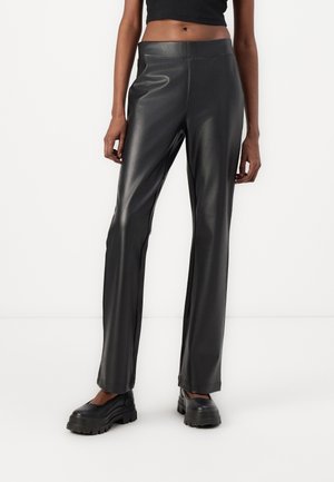 Frau trägt schwarze, hoch taillierte, gerade geschnittene Hose aus Kunstleder mit schwarzen, klobigen Plateau-Schuhen, steht vor weißem Hintergrund.