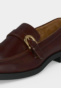 Mocassin en cuir marron avec détail de boucle dorée et semelle noire, présentant une finition lisse et une doublure intérieure matelassée.
