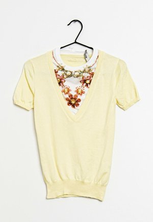 Lichtgele trui met korte mouwen en diepe V-hals, gelaagd over een wit overhemd met een print van een rood en geel bloemenparelketting, aan een zwarte hanger.