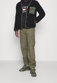 Černá fleece bunda s zelenými akcenty a patentním uzávěrem, spárovaná s olivovými cargo kalhotami. Zeleno-černé tenisky dotvářejí outfit.