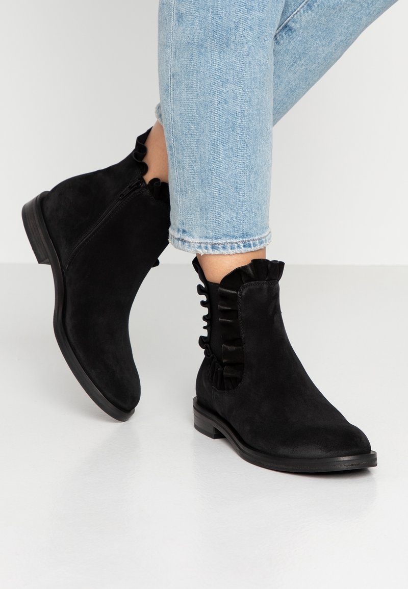 Bottines en daim noir avec bords froncés portées avec un jean bleu clair retroussé, présentées sur un fond de sol blanc.