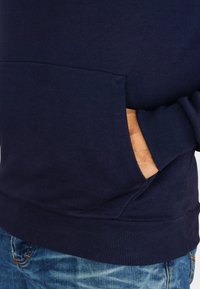 Marineblauer Hoodie aus weicher Baumwolle mit einer frontalen Kängurutasche, gerippten Bündchen und Saum. Jeans mit leichter Abnutzung, die teilweise sichtbar ist.