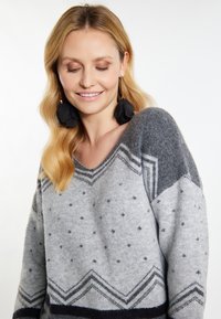 Grauer Strickpullover mit V-Ausschnitt, geometrischen Mustern und unterschiedlichen Texturen. Akzente setzen dunkelgraue und marineblaue Details.