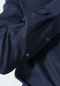 Maglietta a maniche lunghe blu navy realizzata in tessuto morbido, con un accento metallico esagonale al polsino e chiusura con bottoni classica.