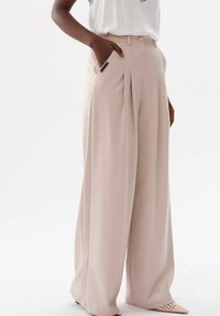 Szerokie, jasnobeżowe spodnie z centralnymi zakładkami, kieszeniami bocznymi i gładką teksturą. Noszone z białą bluzką i jasnym obuwiem.